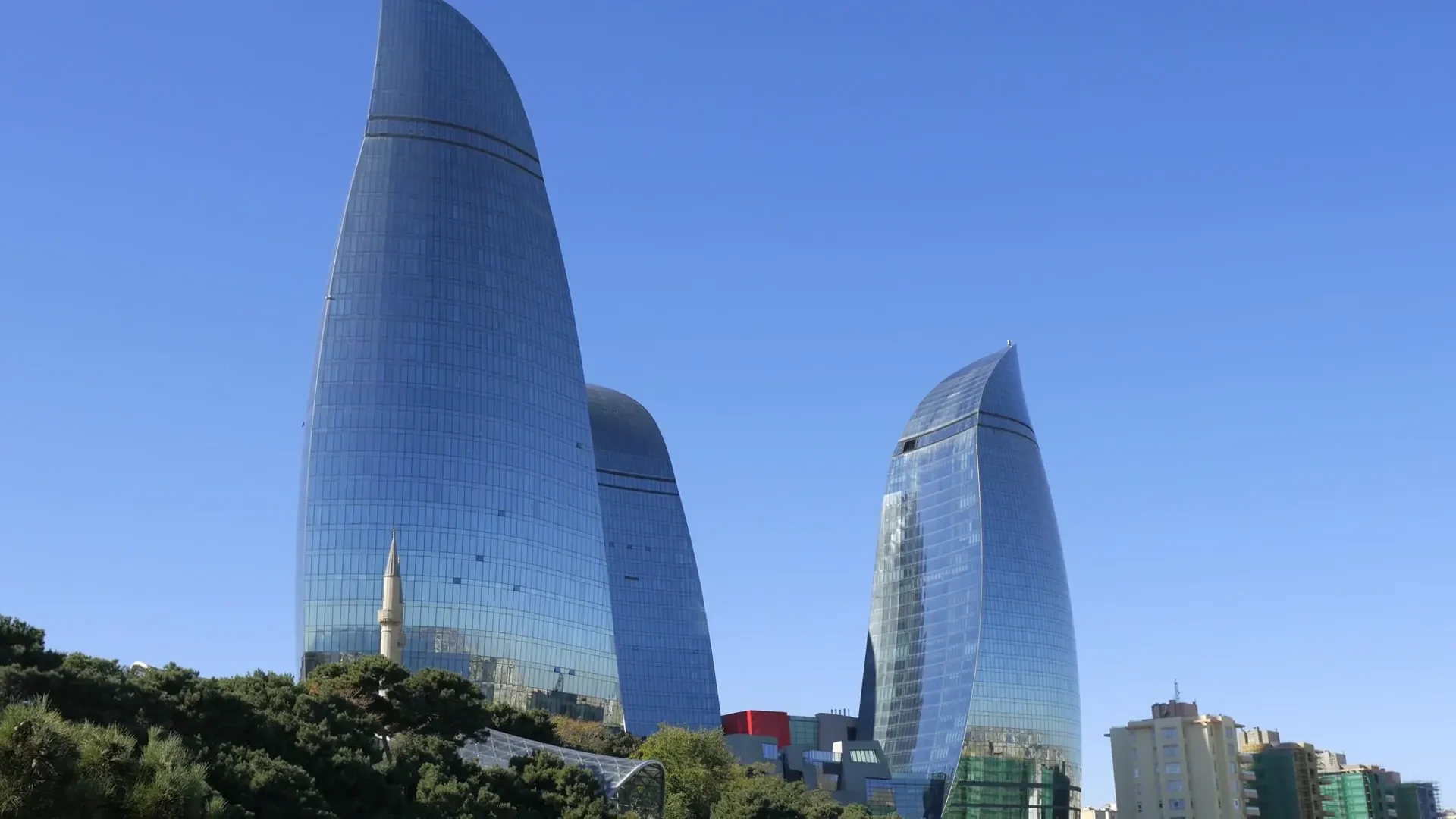 The Flame Towers above the city, Baku, Azerbaijan | Karobar Gateways The Flame Towers rising behind a mosque minaret and a green hillside in Baku